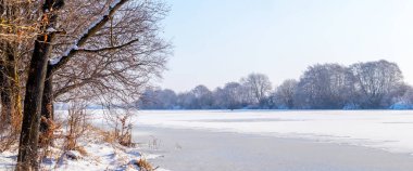 Güneşli bir günde, buzla kaplı bir nehrin yakınındaki ormanın olduğu kış manzarası.