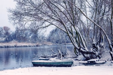 Önünde bir tekne ve arkasında karla kaplı ağaçlarla karla kaplı bir kış gölü kıyısı sakin bir panoramik kış manzarası yaratıyor.
