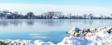 Donmuş gölü, karla kaplı kıyıları ve parlak kış güneşi altında ufukta ağaçlarla panoramik kış manzarası.
