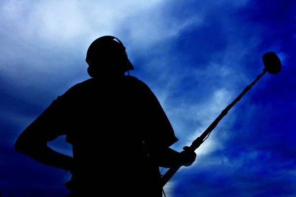 Silhouette of unrecognizable sound technician holding boom microphone isolated over dramatic blue sky.