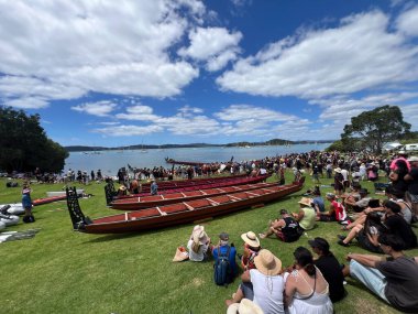 WAITANGI, NZL - FEB 06 2025:Crowd of people sit around war canoes on Waitangi Treati Ground on  Waitangi Day, the national day of New Zealand, marks the anniversary of the 1840 Treaty of Waitangi clipart