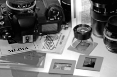 Profesyonel bir kamera, fotoğraf objektifleri ve arka plandaki slaytlarda bir hoparlör yanında medya ve basın kartı bulunan foto muhabiri masası