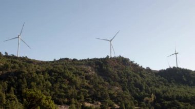 Elektrik Rüzgarı Türbinleri Yeşil Çam Ağaçlarıyla kaplı Dağ yamacında çalışıyor.