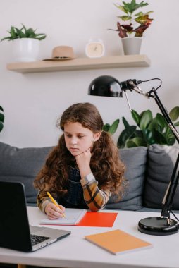 Küçük şirin bir kızın notlar yazarken, dizüstü bilgisayar ve kitaplarla çalışırken, evde okul ödevleri yaparken, notlar yazarken, konferans veya müzik dinlerken, uzaktan eğitim konsepti okurken dikey çekimi.