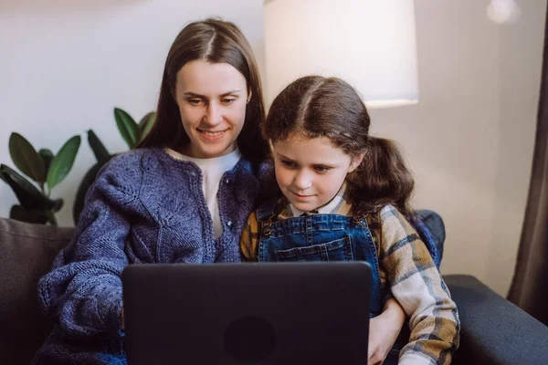 Selective focus of positive young mother with pretty little daughter using laptop, happy mum and cute girl child sitting on sofa in living room at home, watching cartoons online or play video game