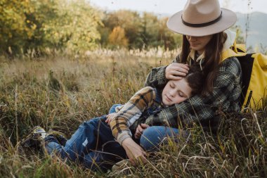 Sonbahar dağlarında çimlerin üzerinde oturan şapkalı ve kızlı bir anne. Seyahat konsepti. Tatil, gezi ve tatil. Dünya Turizm Günü. Güneşin batışında yürümekten zevk alan bir çocuk. Etkin açık hava oyunları