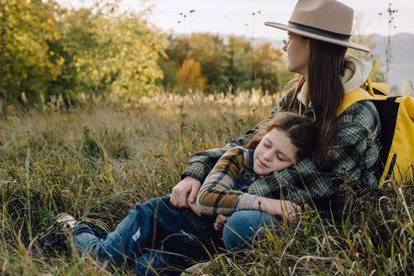 Calm young mother and adorable little daughter lying in grass on background epic autumn mountains, enjoying beautiful nature during sunset. Family trip on fall vacation. Holiday and travel concept
