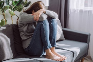 Mutsuz, kaygılı, yüzü diziyle örtülen beyaz bir kadın. Oturma odasında kanepede oturuyor. Kalbi kırık bir kadın depresyonda.