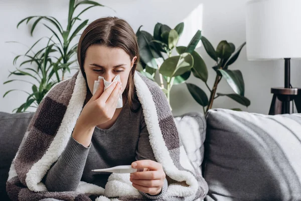 Ill young brunette woman have cold and high fever while checking body temperature by using digital thermometer. Sick girl covered plaid sitting on couch at home. Daily lifestyle healthcare concept