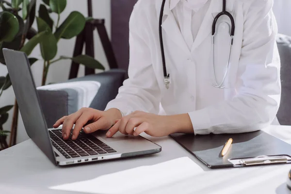 stock image Close up of professional young woman doctor in white coat with stethoscope using laptop sitting on comfy couch at desk in clinic, nurse working on computer at room office. Medical work concept