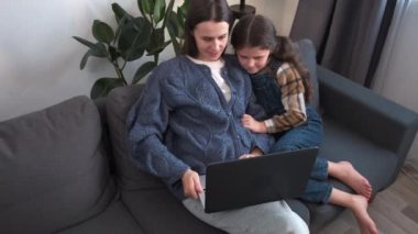 Rahatlamış genç bir anne ve sevimli küçük kızı dizüstü bilgisayar kullanıyor, mutlu anne ve kız çocuğu evdeki rahat koltukta oturuyor, internetten çizgi film izliyor ya da video oyunu oynuyor, aile eğleniyor.
