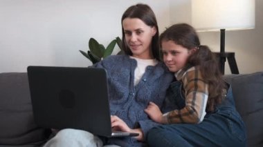 Gülümseyen genç bir anne, sevimli küçük kızına dizüstü bilgisayar kullanmayı öğretiyor. Oturma odasındaki rahat koltuğa oturuyor, mutlu anne ve güzel kız eğleniyor, video izliyor, uygulamaları tarıyor.