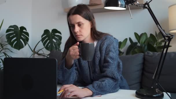 真剣な女の子はカップコーヒーを保持し ブログを書くか ソーシャルネットワークで友人とチャットします ノートパソコンで仕事をしている女性ジャーナリストで自宅のソファに座って画面を見ています — ストック動画
