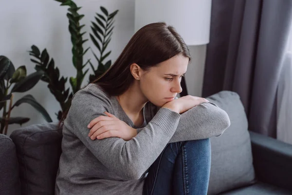 Close up of sad pensive young caucasian woman looking down depressed sitting on sofa in living room at home, thinking about personal problem, break up with boyfriend or divorce. Feeling lonely concept