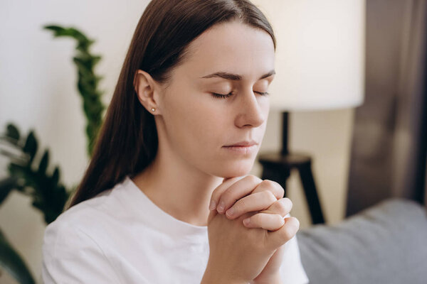 Cute girl praying sincerely with folded arms and making wish, asking with hopeful imploring expression, begging apology. Calm young woman sitting on couch at home living room. Please, God help.