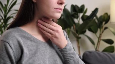 Kış konseptinde grip mevsiminin belirtileri. Evdeki kanepede oturan, boynuna dokunan, boğaz ağrısı, anjin, yutkunması zor, sesi kısılmış, sağlıksız genç beyaz kadının yakın çekimi.