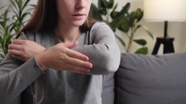 肘に痛みがある若い女性は 外傷後に負傷した手を暖める 悲しい女性のソファマッサージ痛みを伴う手に座っている 肘の痛みに苦しんで彼女の肘を保持する不健康な女の子 選択的焦点 — ストック動画