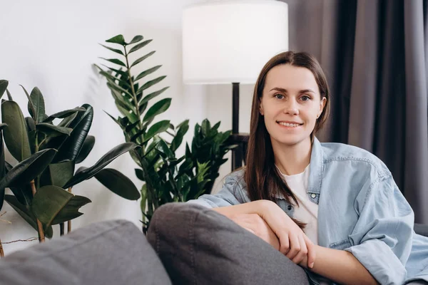 stock image Happy young woman sitting on couch at home, smiling looking at camera. Portrait of adorable girl 20s old years relaxing on sofa. Cheerful beautiful brunette female and relaxing during autumn or winter