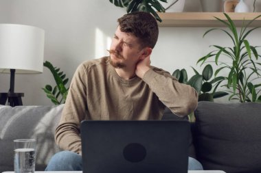 Yorgun ve hasta adam bilgisayarla boğuşuyor sabit yaşam tarzı sırt ağrısı ya da ağrısıyla boğuşuyor. Masa başındaki koltukta oturan yorgun ve sakallı erkek boyun kasları kasılmış.