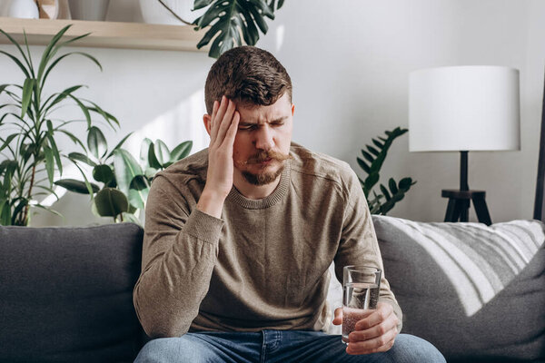 Upset young caucasian man suffering from strong headache or migraine sitting on sofa at home with glass of water, guy feeling intoxication and pain touching aching head. Morning after hangover concept