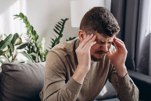 Close up of stressed sad young man feel pain having terrible strong headache concept, tired upset caucasian guy massaging temples suffering from migraine sitting on couch in living room at home