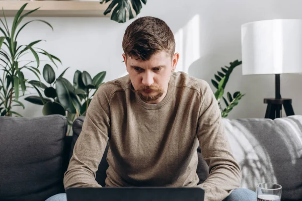 stock image Close up of focused young man sitting on couch with laptop, working remotely online from home. Concentrated millennial male studying distantly alone in living room at home. Freelance concept