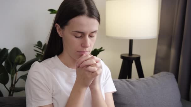 对上帝的信仰 宗教概念 安详的年轻女子紧闭双眼坐在沙发上 双手叉腰祈祷 诚挚地祈求奇迹 祈求好运 表达内心的感激之情 — 图库视频影像