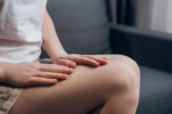 Close-up of female legs with fatigue and cramp. Legs pain concept. Unrecognizable unhealthy young woman with quadriceps pain, feels discomfort, does massage itself sitting on couch at home
