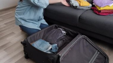 Gülümseyen genç bayan seyahat için hazırlanıyor, büyük bavulda giysilerini topluyor. Seyahat kavramı, macera. Güzel bir milenyum dişisi sıcak zeminde oturur ve yolculuktan önce en gerekli şeyleri toplar.