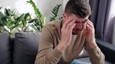 Stresli genç bir adamın yan görüntüsü 30 'lu yaşlarda acı çekiyor korkunç bir baş ağrısı konsepti var, yorgun sakallı adam migrenden muzdarip şakaklara masaj yapıyor evde oturma odasındaki gri kanepede oturuyor.