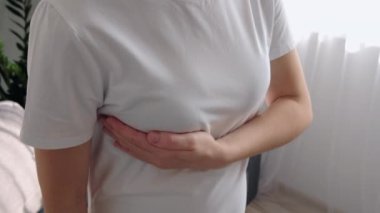 Sağlıksız, beyaz kadın göğsüne dokunuyor. Evinin penceresinin yanında duruyor. Hasta kız beyaz tişört giyiyor. Göğüs kanseri kontrolü. Sağlık ve Tıp Konsepti