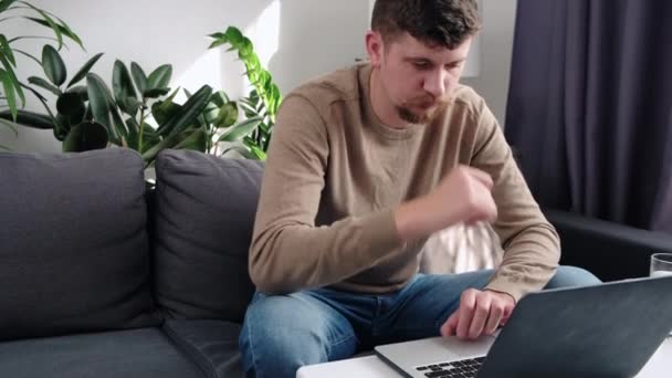 ノートパソコンのガジェットで自宅の仕事でソファに座っている焦点を当てた若い白人男性のクローズアップ コンピュータ上の集中型ひげそりの男性の入力は デバイス上の無線インターネットを参照してください 技術コンセプト — ストック動画