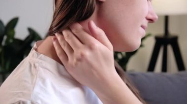 Boyun ağrısı, boyun ağrısı konsepti. Kafkasyalı üzgün genç bir kadının boynuna dokunup yoğurması, evdeki kanepede oturmaktan rahatsızlık duyması. Boyun ağrısı olan acı çeken bir kadının yüzünü yakından çek.