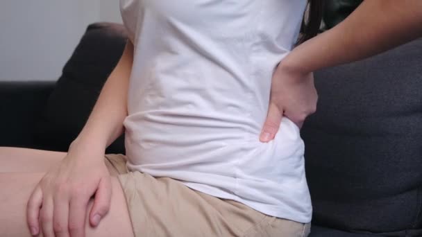 近距离观察患有背痛 骨质疏松症的年轻女性 用手触摸背部 坐在家中灰色的沙发上 按摩较低 保健和医疗概念 — 图库视频影像