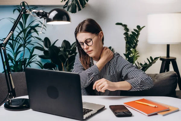 Tired young brunette woman in eyeglasses with pain neckache works at computer sitting on couch in home office at night. Neck pain, pinched nerves, tensed sore muscles, cervical osteochondrosis concept