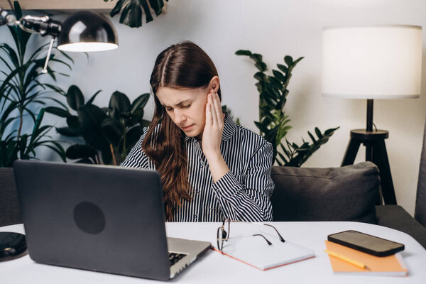 Upset young brunette female sit on sofa at table working laptop feeling ear pain, unhappy irritated businesswoman or student. Furuncle, earache, shingles, eczema, ear disease, otitis concept
