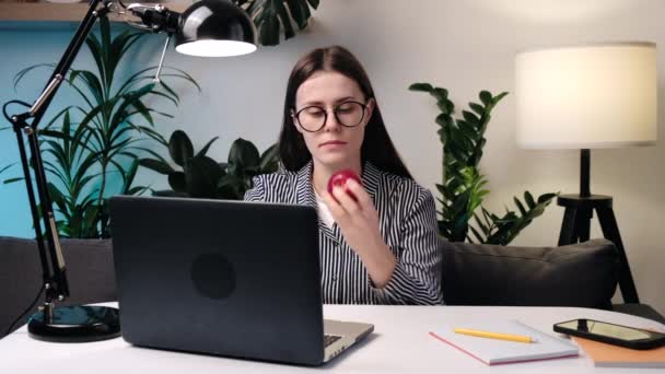 快乐的年轻女人戴着眼镜 坐在沙发上吃苹果 女自由职业者在电脑上观看在线学习 可爱的女孩在家里坐在笔记本电脑前 吃零食 健康饮食 — 图库视频影像