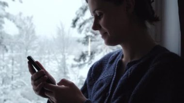 Çekici genç bir kadının, rahat pencere eşiğinde oturduğu ve elinde cep telefonuyla güzel kış manzarasının tadını çıkardığı yakın profil. Akıllı telefon başvuru pazarı, podcast dinleme. Rahatlama kavramı