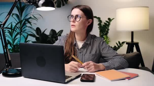 眼鏡をかけペンを保持している若い女性は 思慮深く見てノートパソコンや個人的な日記とソファに座っている間に距離を見つめてアイデアを考えます 検索ソリューションと創造的な意思決定 — ストック動画