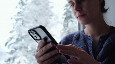 Camın kenarında oturan ve elinde cep telefonuyla güzel kış havasının tadını çıkaran çekici genç bir kadının seçici odağı. Akıllı telefon başvuru pazarı, podcast dinleme. Rahatlama kavramı