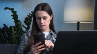 Dizüstü bilgisayarı olan endişeli genç bir kadın evdeki kanepede oturan kafası karışık bir şekilde cep telefonuna bakıyor. Beklenmedik mesajları okuyan stresli bir bayan, cep telefonundan üzgün bir e-posta aldı.