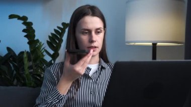 Ciddi genç iş kadını evdeki bilgisayarda çalışıyor. Kız akıllı telefondan sesli mesaj kaydediyor. Odaklanmış Kafkas kadın çalışanı cep telefonundaki hoparlörde dizüstü bilgisayarla alışveriş yapıyor.