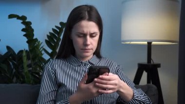 Stresli, endişeli, kıskanç, cep telefonlu, üzgün bir kız. Kanepede yalnız otururken mesaj almanın sorun olacağını düşünüyor. Endişeli genç bayan evde akıllı telefondan kötü haber okuyor.