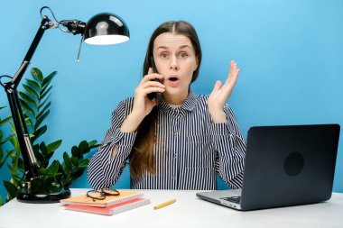 Beyaz ofis masasında çalışan kafası karışık bir çalışan portresi cep telefonuyla konuşan cep telefonuyla konuşan, stüdyoda mavi renkli arka plan duvarına izole edilmiş bir poz veren.