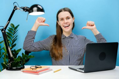 Mutlu, kendine güvenen başarılı, gömlekli iş kadını beyaz ofis masasında çalışıyor bilgisayarın işaret parmağıyla kendini işaret ediyor, stüdyonun mavi arka plan duvarına izole edilmiş pozlar veriyor.