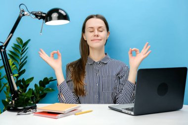 Başarılı çalışanın portresi beyaz ofis masasında dizüstü bilgisayarla yoga yaparken el ele tutuşup rahatla meditasyon yapmaya çalış, sakin ol, mavi arkaplan duvarının arkasında izole bir şekilde poz ver.