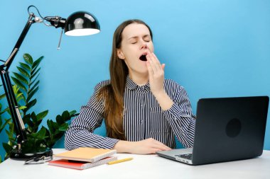 Yorgun, uykulu, yorgun, beyaz gömlekli genç bir kadının portresi ofis masasında bilgisayarla çalışıyor esniyor, stüdyodaki pastel mavi arka plan duvarında izole bir şekilde poz veriyor.