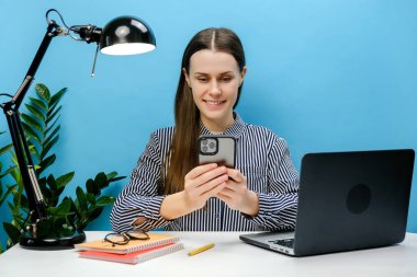 Gülümseyen başarılı bir çalışan genç bayan cep telefonuyla beyaz ofis masasında oturuyor internette geziniyor, mavi renkli arka plan stüdyosuna izole edilmiş pozlar veriyor.