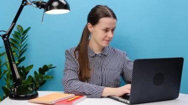 Başarılı çalışan, 20 'li yaşlarda rahat gömlekli genç bir kadın, beyaz ofis masasında laptopla çalışır, kazanan jestler yapar, stüdyodaki pastel mavi arka plan duvarında izole bir şekilde poz verir. Başarı kariyer konsepti