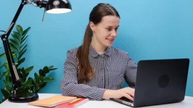 20 'li yaşlarda, rahat gömlekli başarılı bir kadının portresi beyaz ofis masasında dizüstü bilgisayarlı parmaklarıyla oturuyor. Nakit para istediğini gösteriyor. Mavi arka plan stüdyosundan izole edilmiş. İş konsepti